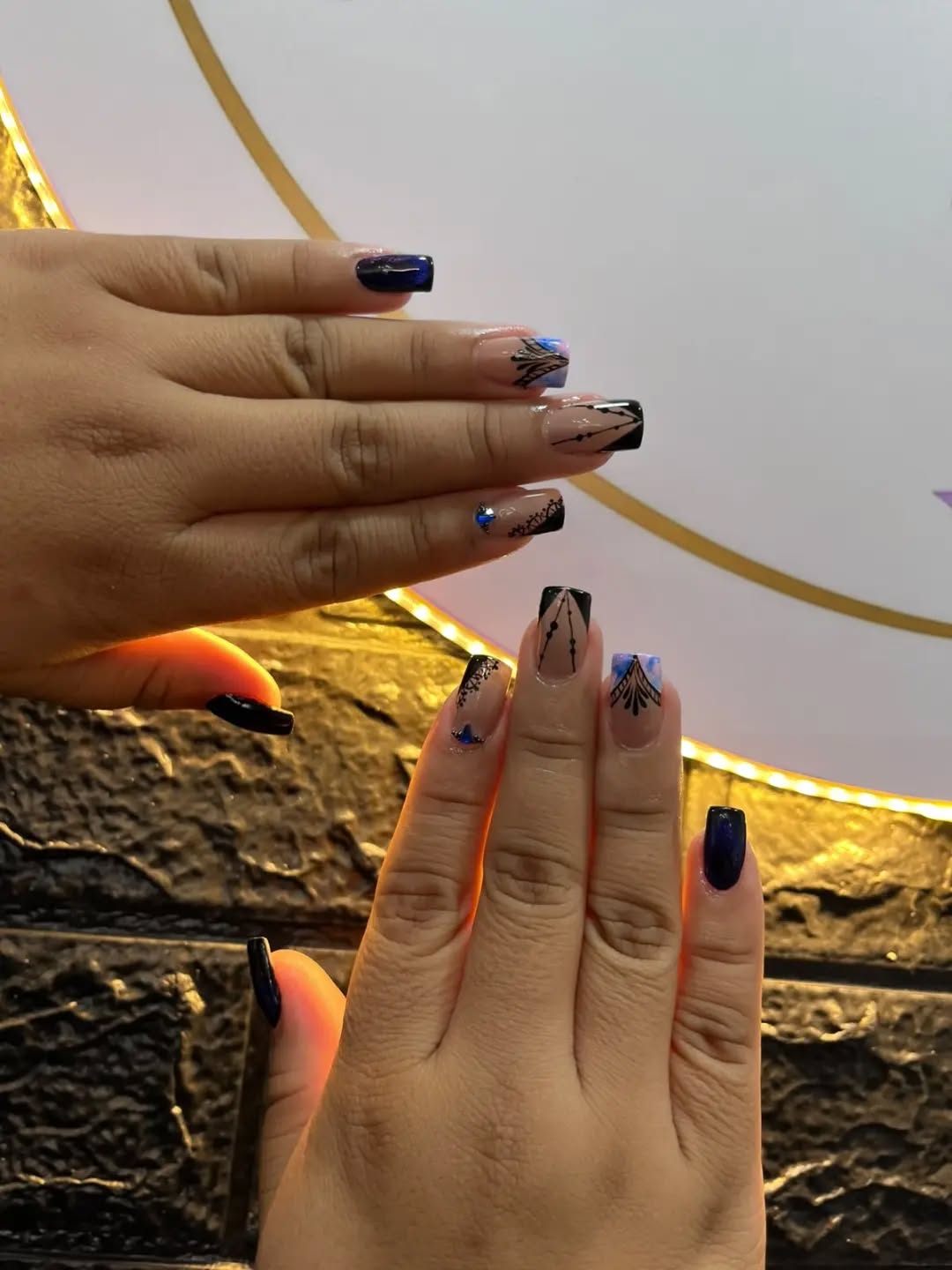 Dos manos con uñas cuadradas en negro, azul y encaje frente a pared dorada