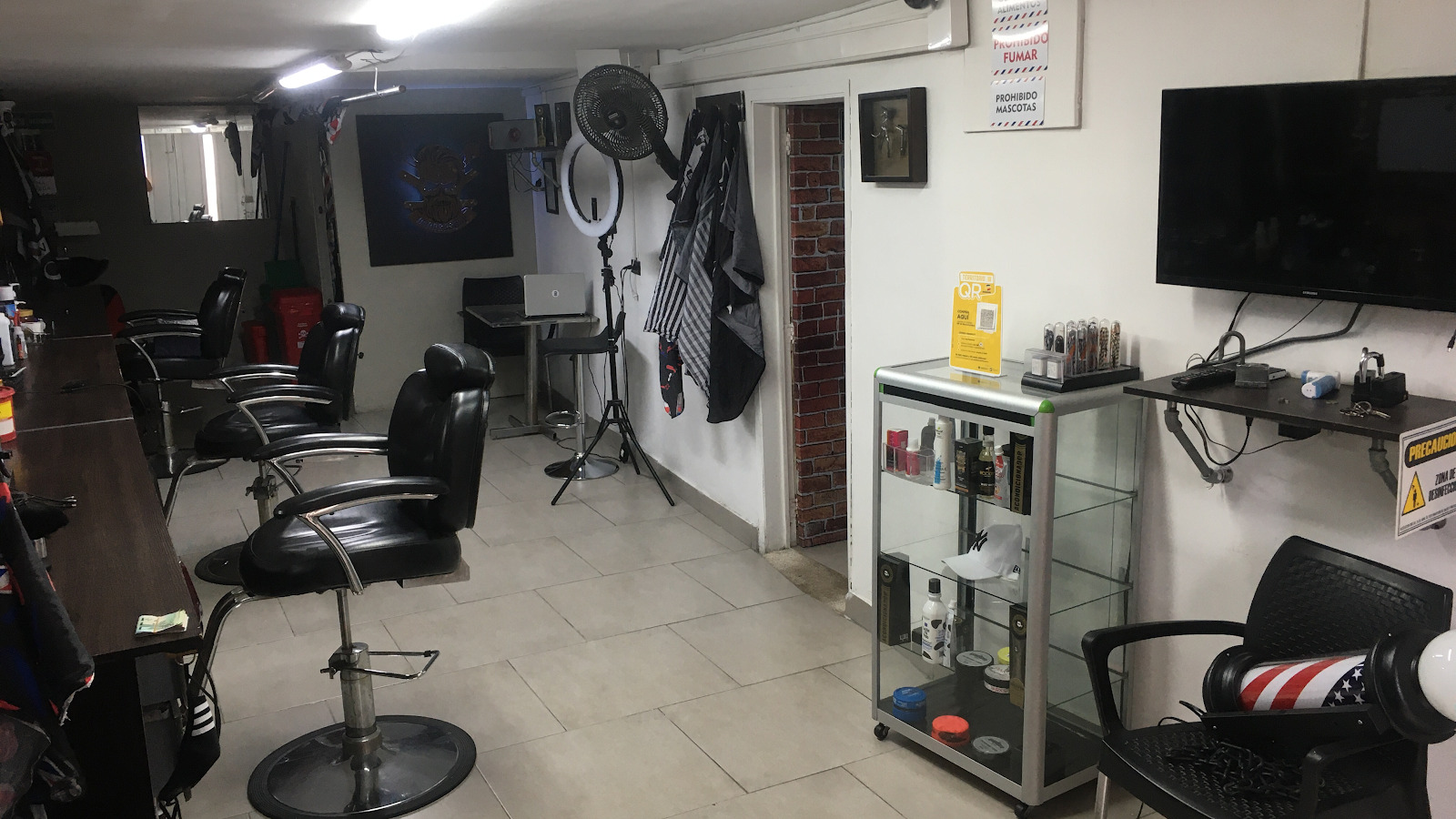 Salón con tres sillas de barbero negras, vitrina de productos, televisor y piso claro